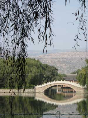 柳葉越しに見える、池と石橋ある中国庭園風の天水馬包泉公園と頂まで耕地化された黄土山地