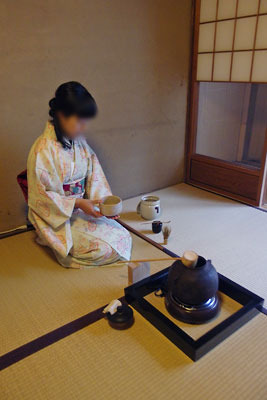 京都の自宅町家で行われた実験茶会で、畳上に置いた電熱や炉縁による代用茶炉の前に座り、薄茶点前を始める亭主役の和装女史
